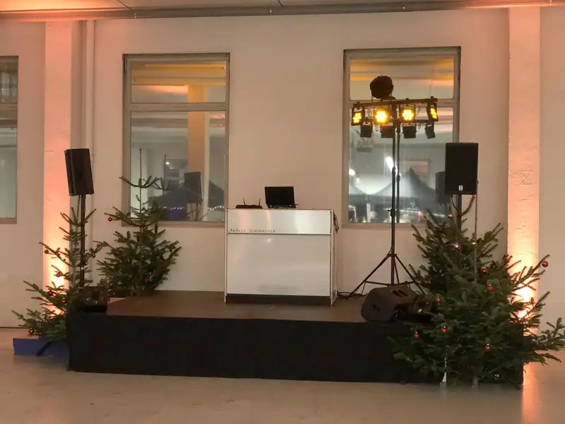 DJ-Tisch von Markus Schumacher mit Lautsprechern und Licht auf einer Bühne in einer Fabrikhalle, geschmückt mit Tannenbäumen für eine Weihnachtsfeier