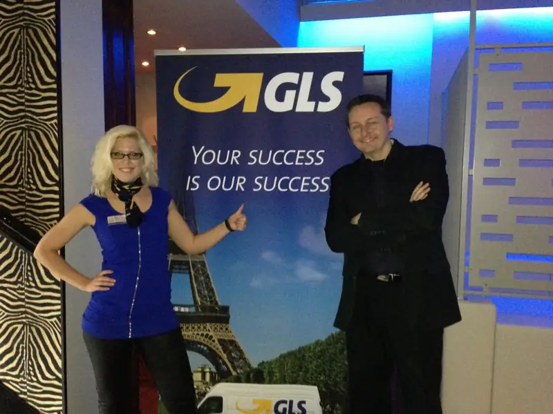 Markus Schumacher steht mit verschränkten Armen neben einem Aufsteller bei einer Firmenveranstaltung von GLS, daneben eine Mitarbeiterin