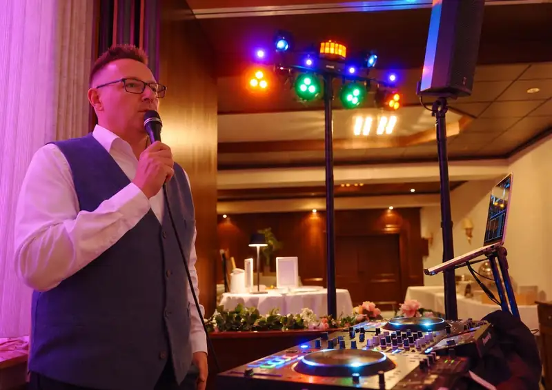 Markus Schumacher moderiert am DJ-Pult, hinter ihm leuchtet die Eurolight LED Bar mit bunten Spots