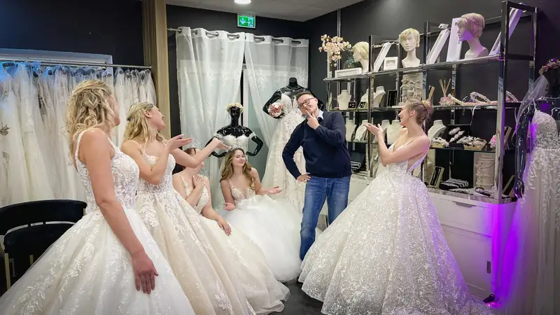 Markus Schumacher steht lachend und nachdenklich mit der Hand am Kinn in einem Brautmodengeschäft zwischen mehreren Bräuten in Hochzeitskleidern