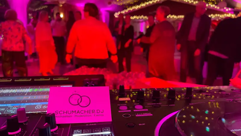 Blick über das DJ-Pult mit der Visitenkarte schumacher.dj auf eine feiernde Gesellschaft im rot-pinken Licht